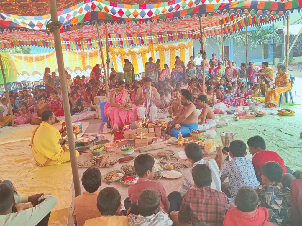 Group of people participating in a traditional ceremony under a colorful canopy, with offerings and decorations present.